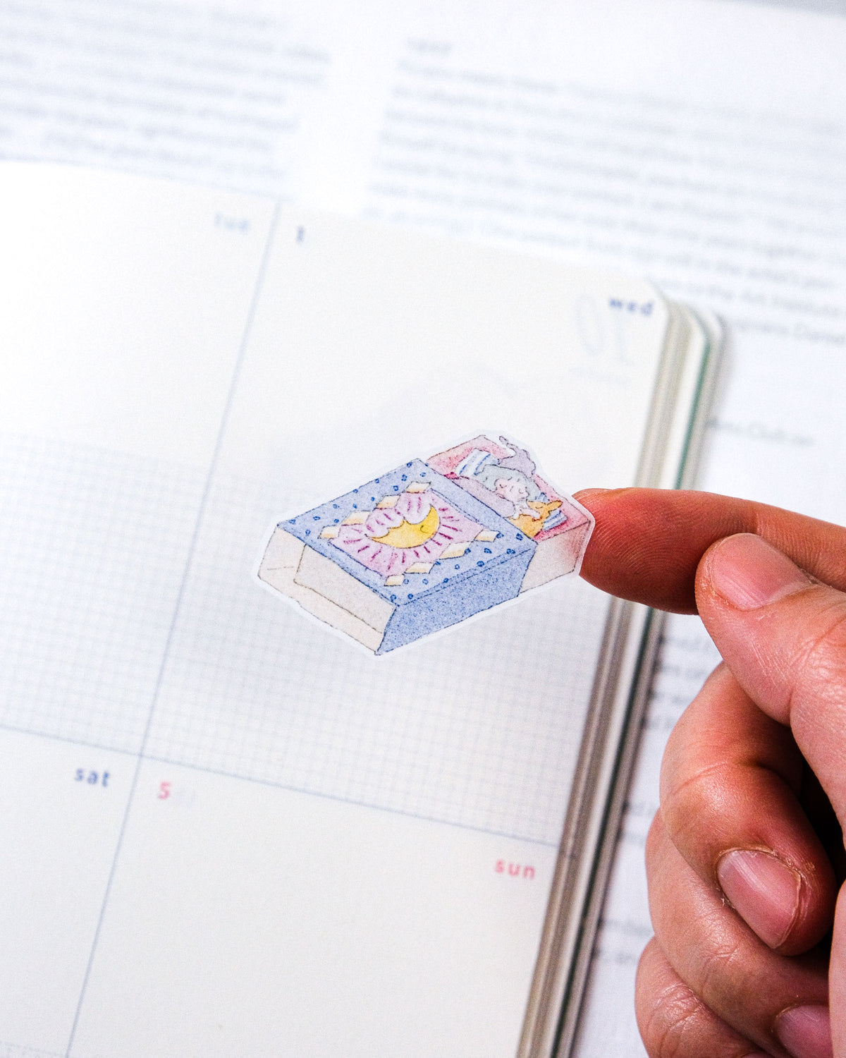 A hand holds a tiny illustrated sticker or charm shaped like a bed with a sun and moon, positioned over an open planner or notebook page.