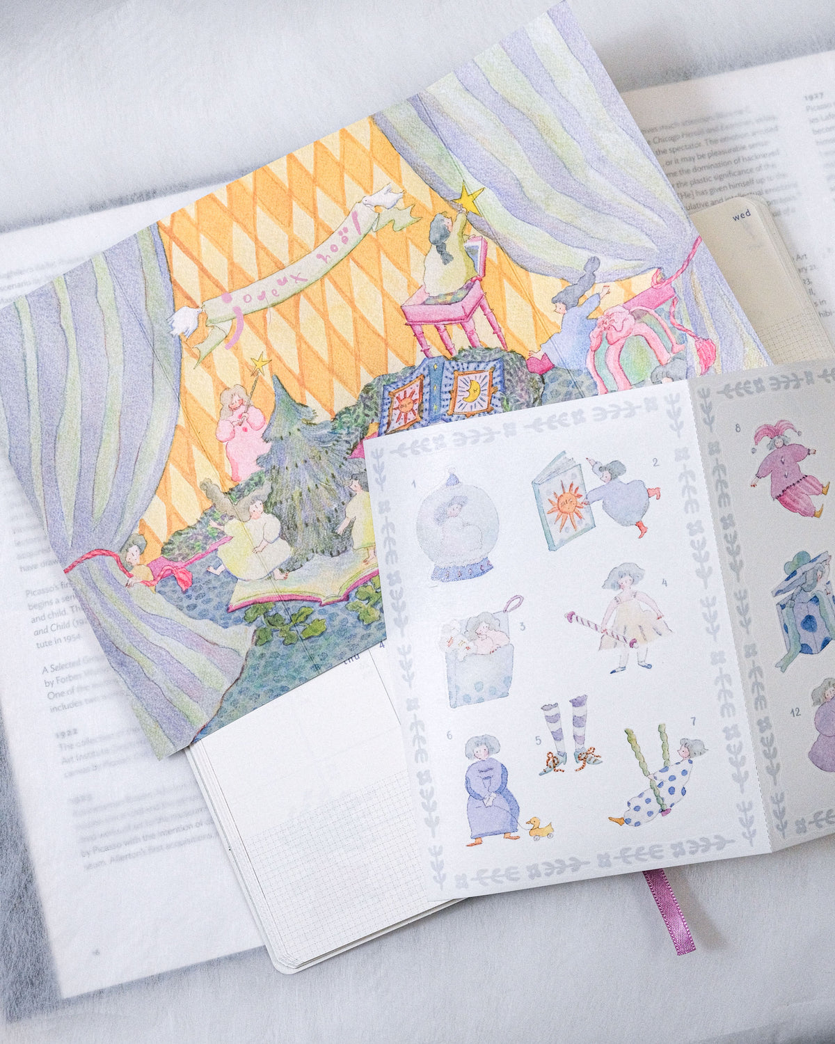 A styled flat-lay of a children's book or sticker album with whimsical illustrated covers and pages, placed on top of open books on a soft gray fabric surface. sticker, christmas, dodolulu
