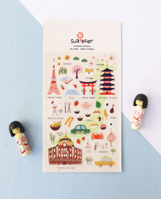 A sheet of Suatelier daily in Tokyo stickers displayed on a light blue and white background, flanked by two small wooden doll figurines. The sticker sheet features various Tokyo-themed illustrations including the Tokyo Tower, Shibuya Crossing, and street food. nz