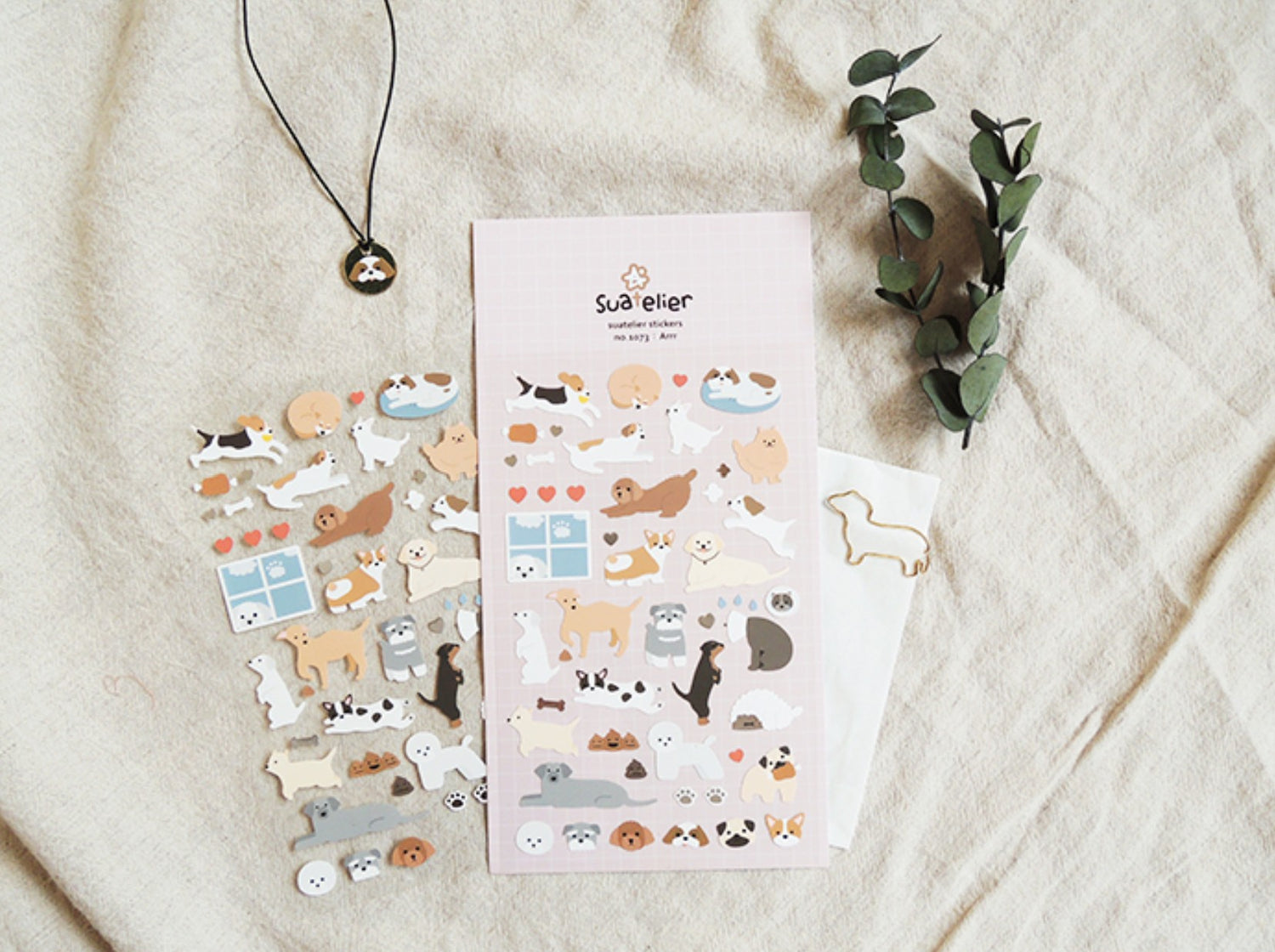 A styled flat-lay of cute dog-themed stickers on a light beige linen fabric background, featuring a pink sticker sheet, a sheet of individual dog stickers, a gold dog-shaped paperclip, and a black pendant necklace with a dog charm. Suatelier, korean, sticker, hope and letters, auckland, nz, dog, puppy