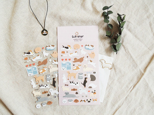 A styled flat-lay of cute dog-themed stickers on a light beige linen fabric background, featuring a pink sticker sheet, a sheet of individual dog stickers, a gold dog-shaped paperclip, and a black pendant necklace with a dog charm. Suatelier, korean, sticker, hope and letters, auckland, nz, dog, puppy