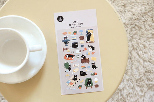 A sheet of cute cat-themed planner stickers displayed on a light beige round table next to a white ceramic cup and saucer, with a soft white textured rug in the background. suatelier, hope and letters, korea, cute, cat, sticker, black cat, nz, auckland