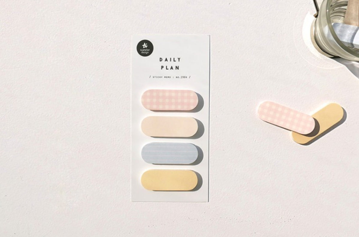A vertical card labeled 'DAILY PLAN 50' with four pastel pill-shaped tabs in pink, cream, light blue, and pale yellow, placed on a white surface beside two loose matching tabs and part of a glass object. suatelier, korea, nz, hope and letters, sticky notes, memo, cute