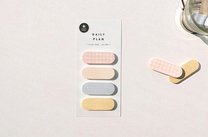 A vertical card labeled 'DAILY PLAN 50' with four pastel pill-shaped tabs in pink, cream, light blue, and pale yellow, placed on a white surface beside two loose matching tabs and part of a glass object. suatelier, korea, nz, hope and letters, sticky notes, memo, cute