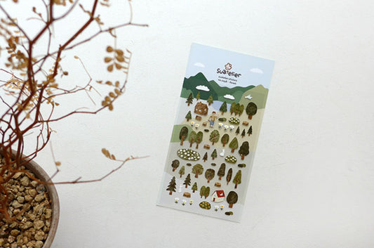 A sheet of Suatelier forest-themed stickers featuring cute woodland illustrations like trees, mushrooms, a squirrel, and mountains, displayed on a clean white surface next to a potted plant. Korean, hope and letters, cute, tree, forest, sticker, nz
