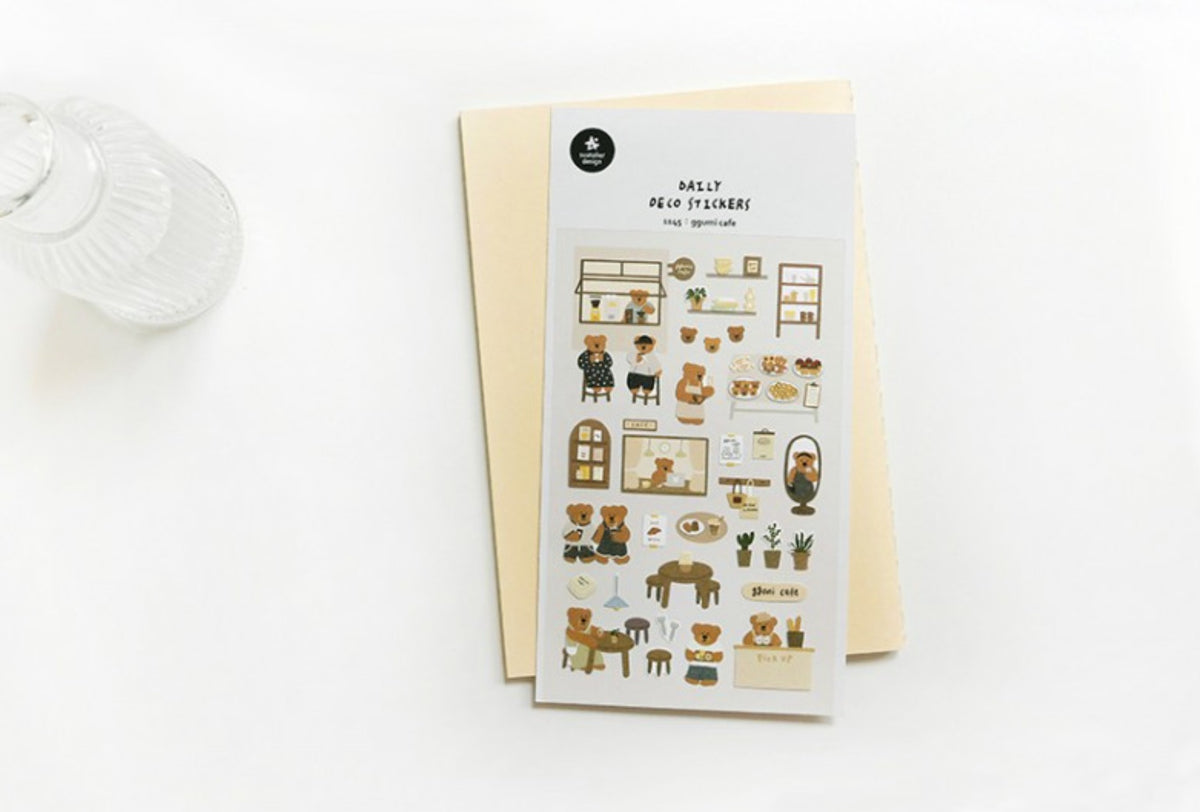 A sheet of mini room-themed stickers titled 'daily deco stickers ggumi cafe' is displayed on top of a cream-colored envelope against a clean white background. The stickers feature cute illustrations of teddy bears, cozy home interiors, and small furniture pieces in soft beige, brown, and black tones.