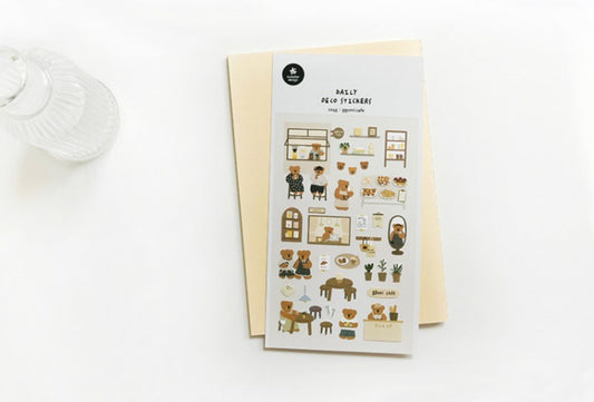 A sheet of mini room-themed stickers titled 'daily deco stickers ggumi cafe' is displayed on top of a cream-colored envelope against a clean white background. The stickers feature cute illustrations of teddy bears, cozy home interiors, and small furniture pieces in soft beige, brown, and black tones.
