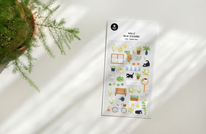 A sheet of cute garden-themed planner stickers displayed on a white surface with soft diagonal shadows, next to a potted green plant. suatelier. nz. cat