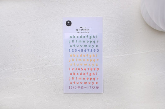 A sheet of alphabet and number stickers in pastel colors, displayed on a textured off-white surface.