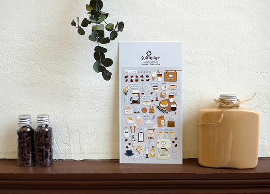 A sheet of coffee-themed stickers by SuZieger stands on a wooden shelf against a textured cream wall, styled with two glass jars of coffee beans and a ceramic bottle on either side.