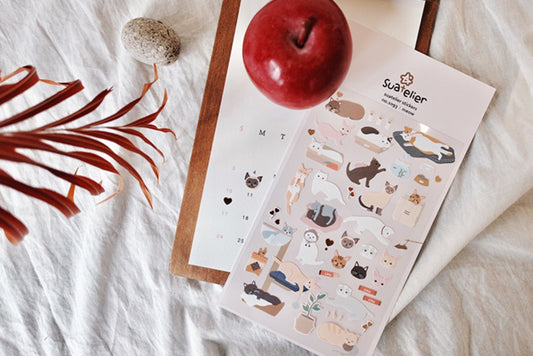 A sheet of cute cat-themed stickers on a light wood tray, styled on a white fabric background with a red apple and dried leaves. suatelier, sticker, cute, cat, pastel, white cat, ragdoll, bengal, nz, hope and letters, auckland